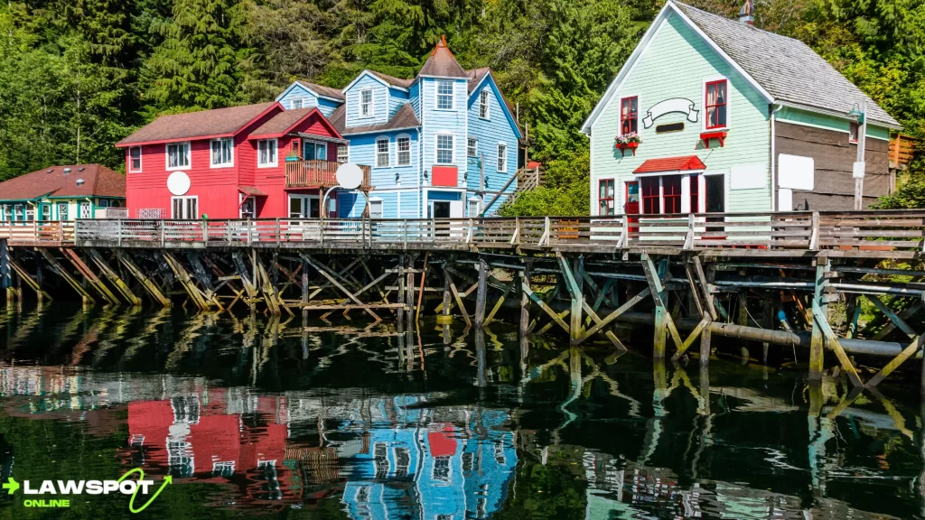 Colorful wooden houses line the waterfront in an Alaskan coastal town, reflecting off the calm water. Can you drive to Alaska? Yes, and picturesque towns like this make the journey even more rewarding.